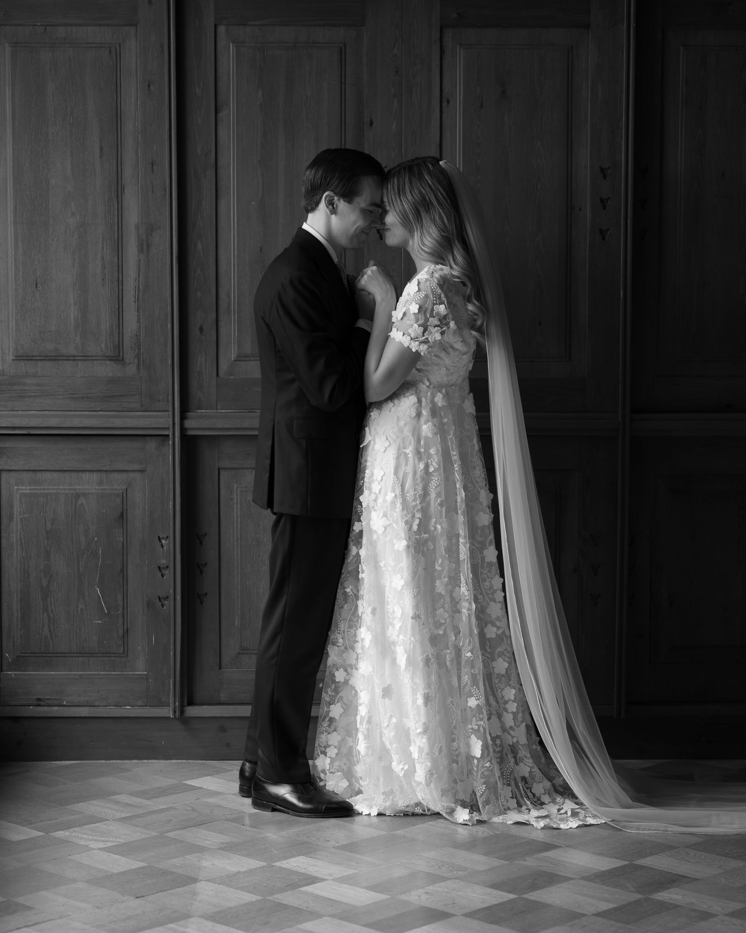 Black and white photo of a bride in a floral lace gown and long veil embracing her groom in a dark suit in a wood-paneled room.