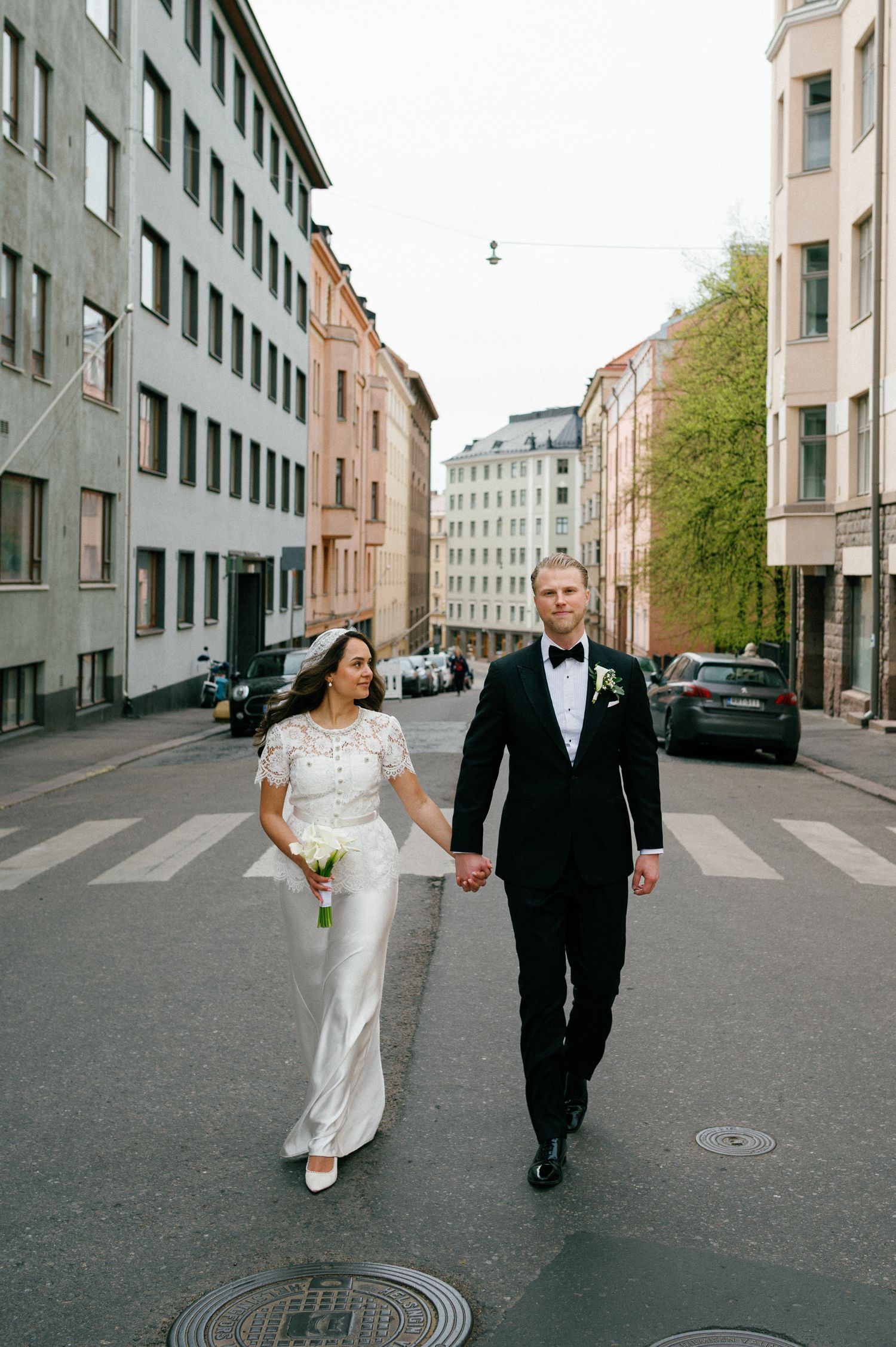 Bride and groom hold hands walking across a city street, she in a white gown, he in a black tuxedo.
