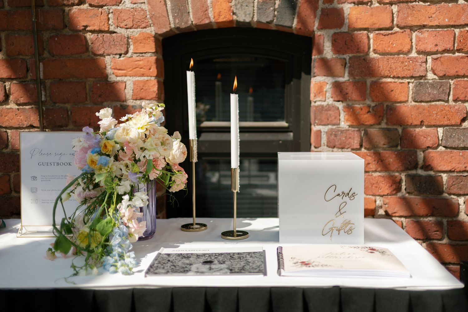 Elegant wedding table with floral arrangement, lit candles in gold holders, ceremony booklet, and personalized white card box.