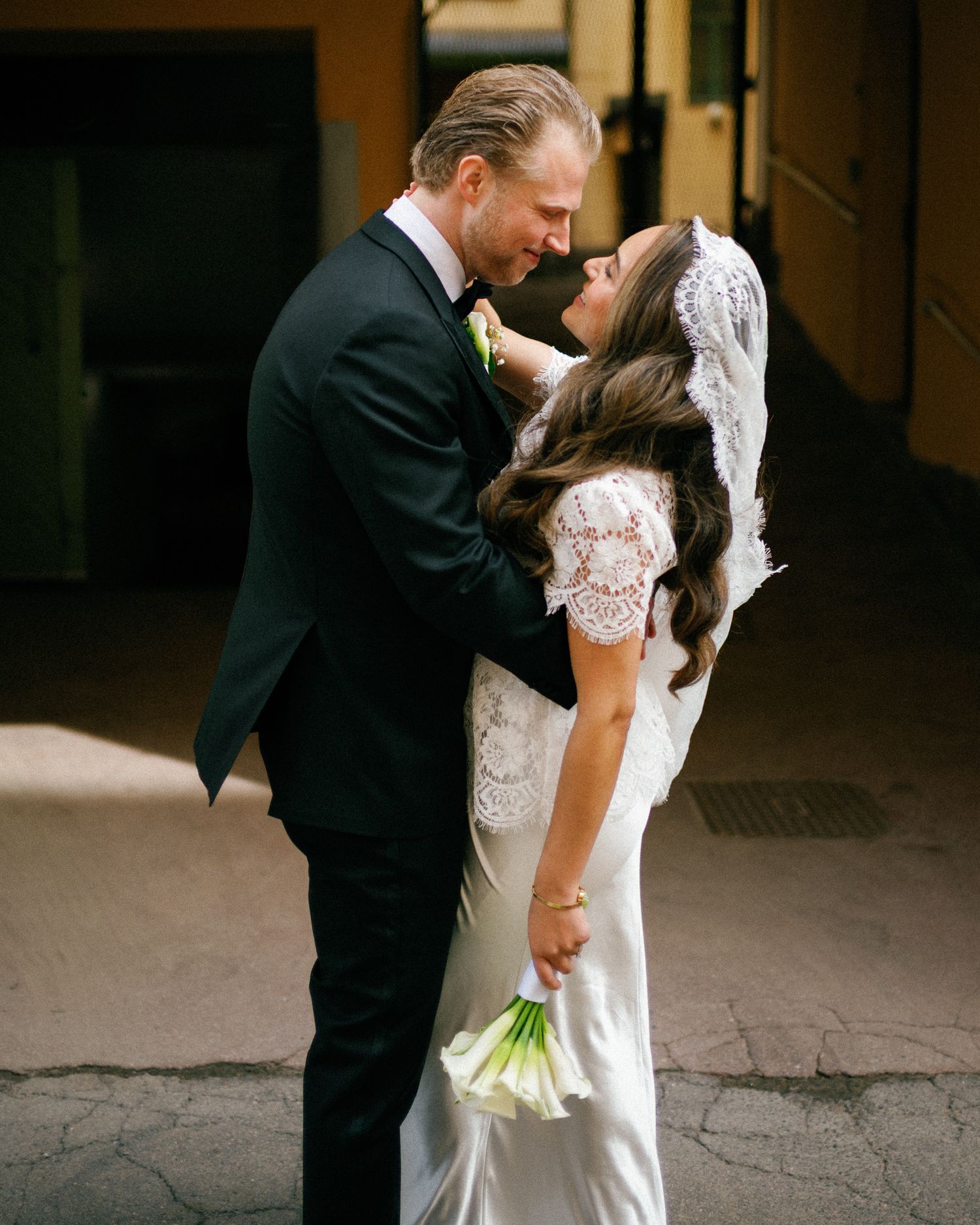 A groom in a dark suit embraces a bride in a lace wedding dress and veil as she holds a white calla lily bouquet.
