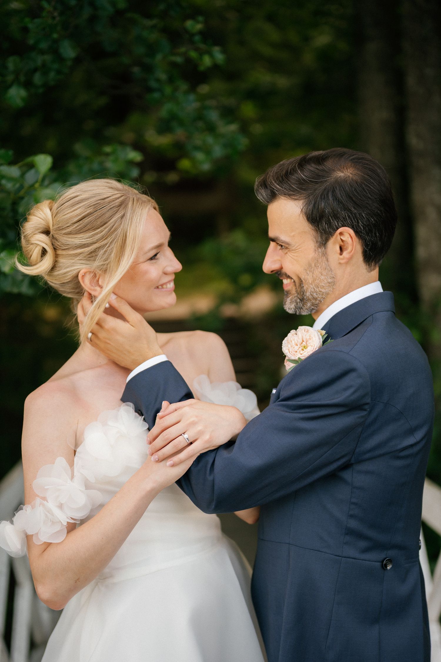 A smiling bride and groom in wedding attire share a romantic moment outdoors surrounded by lush green trees.