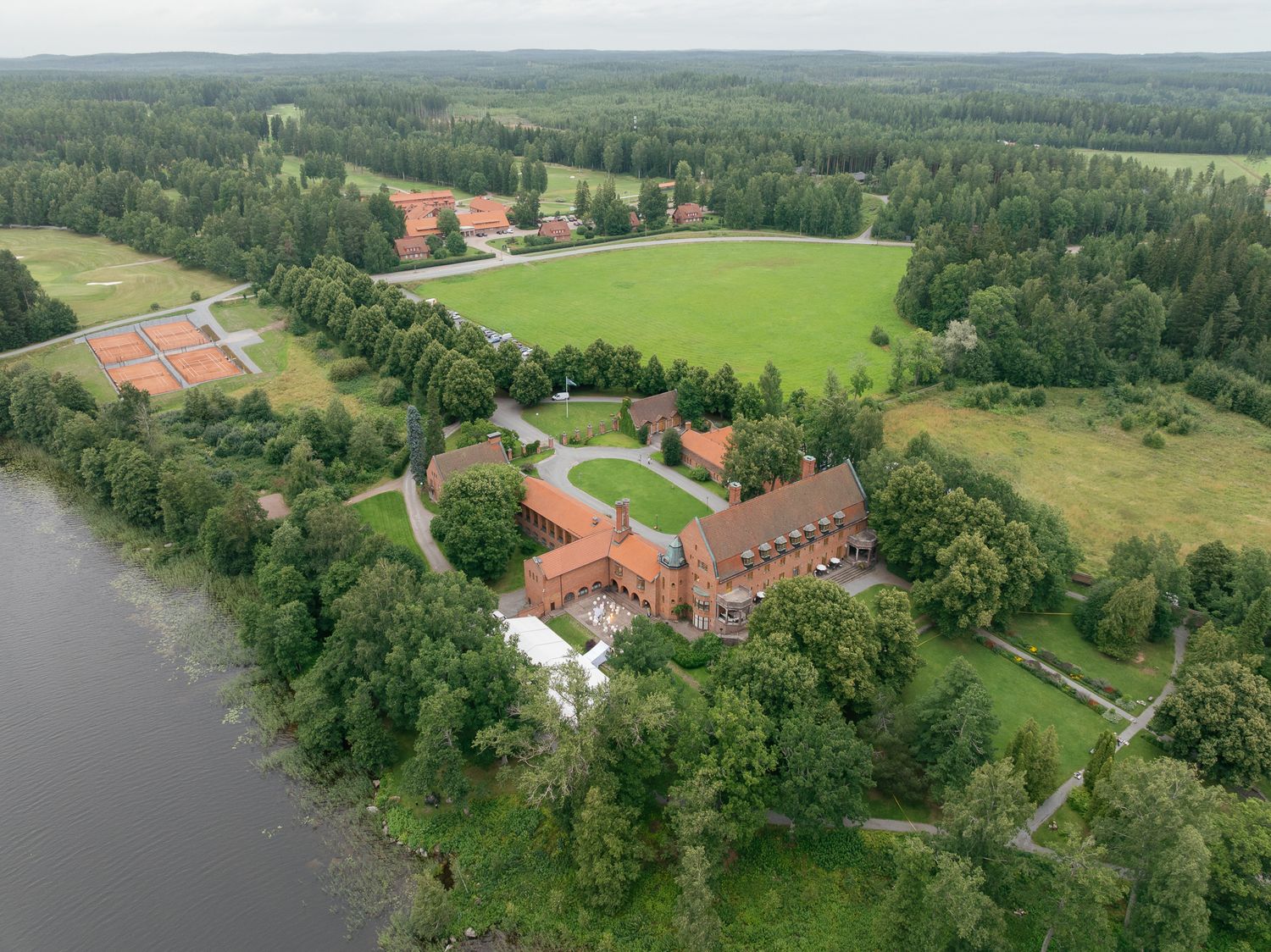 Aerial view of a red brick estate with tennis courts, green fields, and forests beside a calm lake.