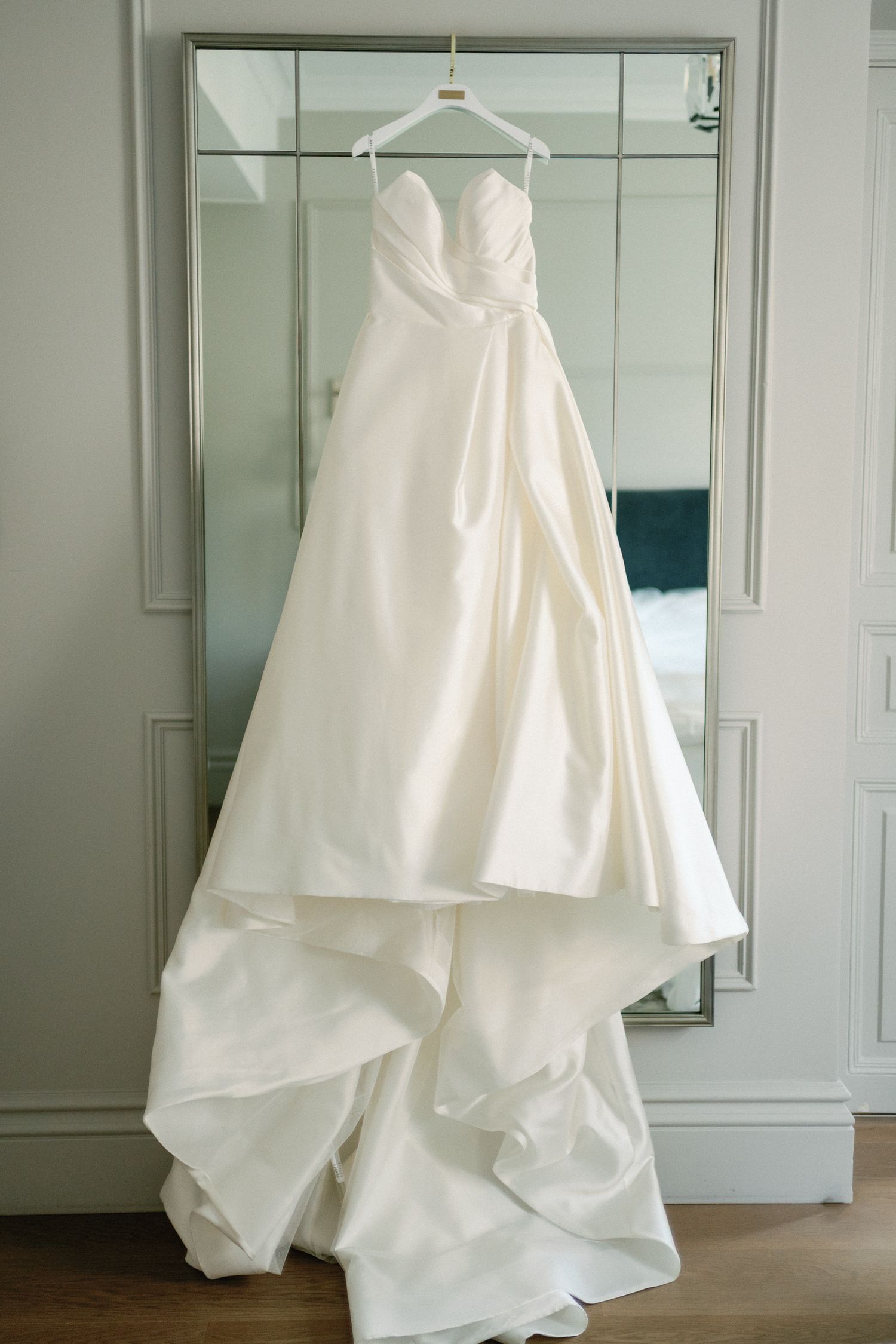 Elegant strapless white satin ball gown wedding dress hanging on a mirror in a bright bridal suite.