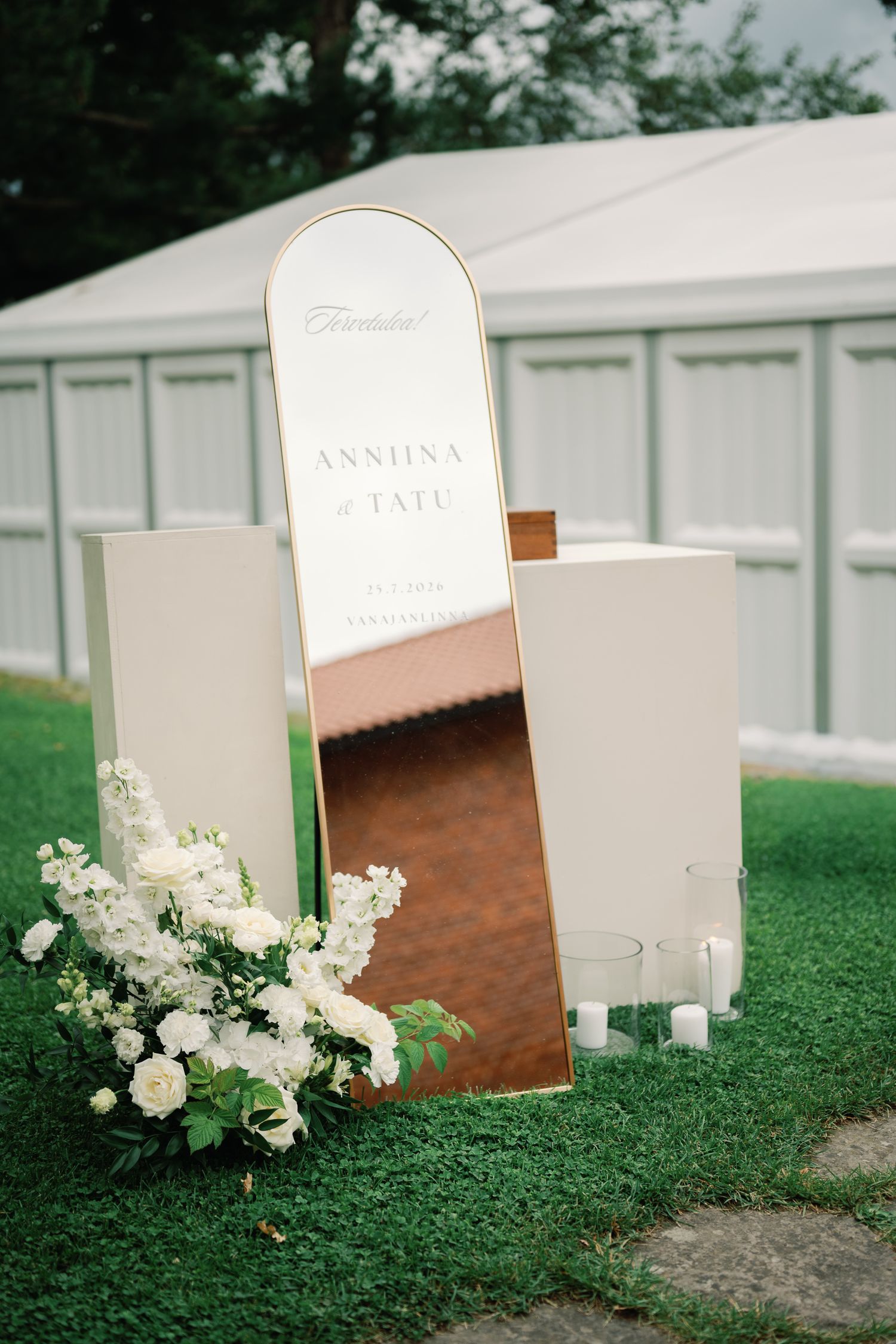 Elegant wedding welcome mirror sign with gold frame, white floral arrangement, and candles on green grass.