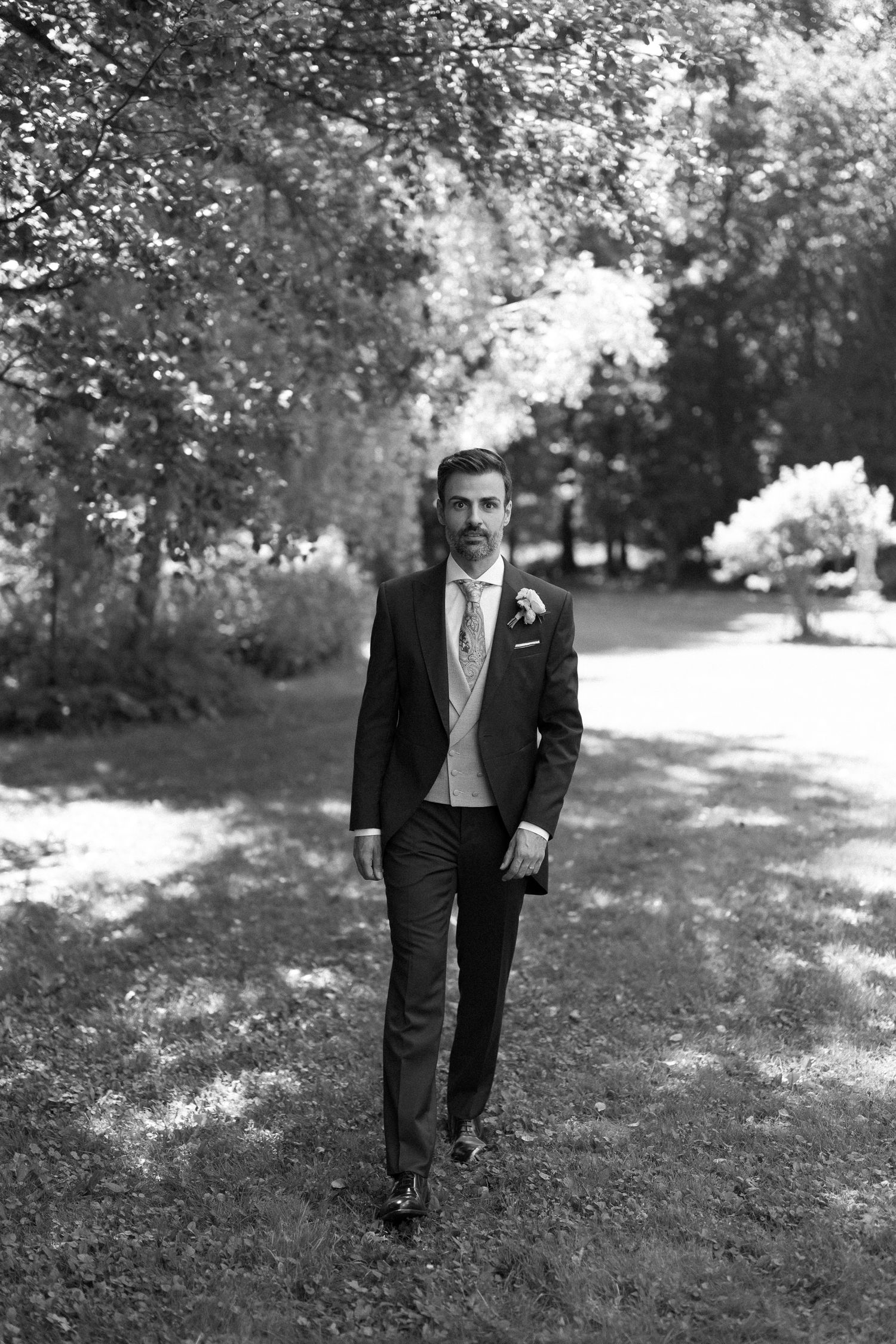 A groom in a dark suit with floral boutonniere walks through a tree-lined garden path in a black and white photo.