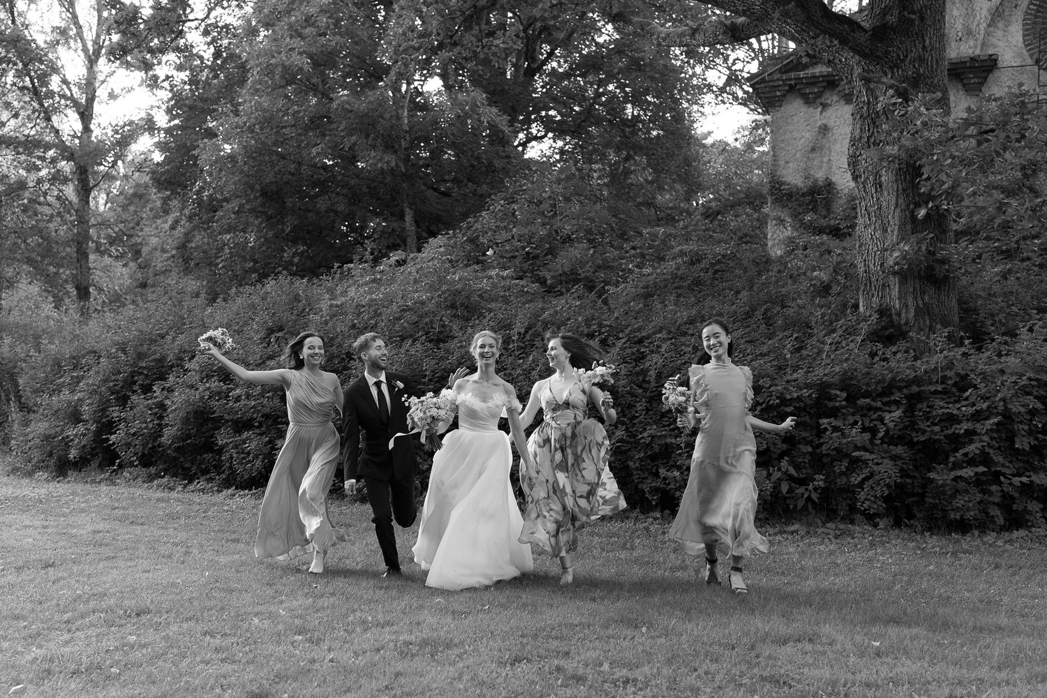 Black and white photo of a bride, groom, and bridesmaids joyfully walking through a lush garden.