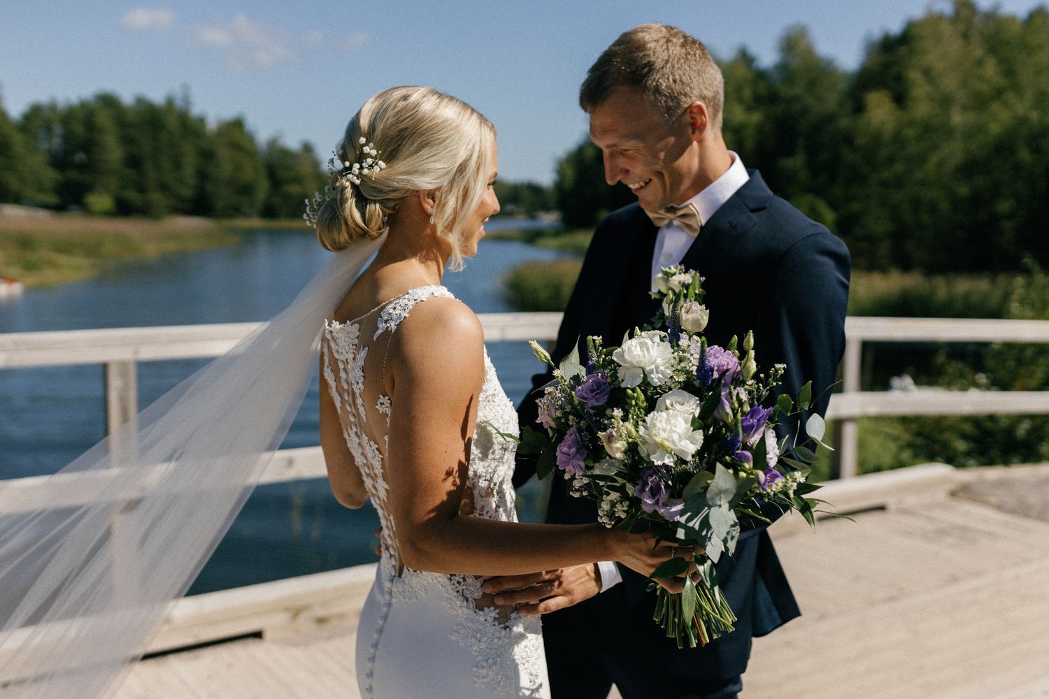 Elegant wedding couple embraces outdoors with a flowing veil and bouquet against a natural backdrop.