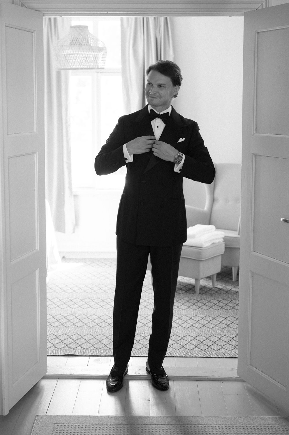 Person in formal black tuxedo adjusting bow tie in white doorway.