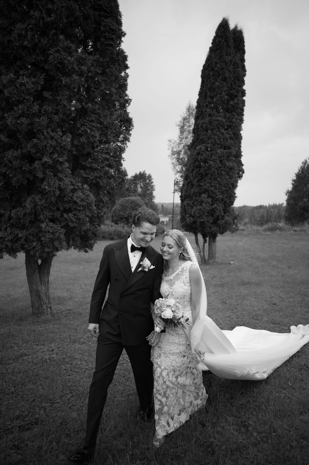 A romantic wedding couple poses together among tall cypress trees in a black and white outdoor portrait.