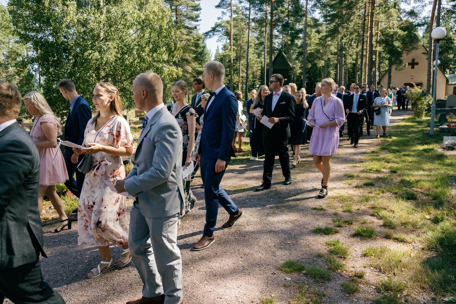 Wedding guests walk along a forest path on a sunny day during an outdoor celebration.