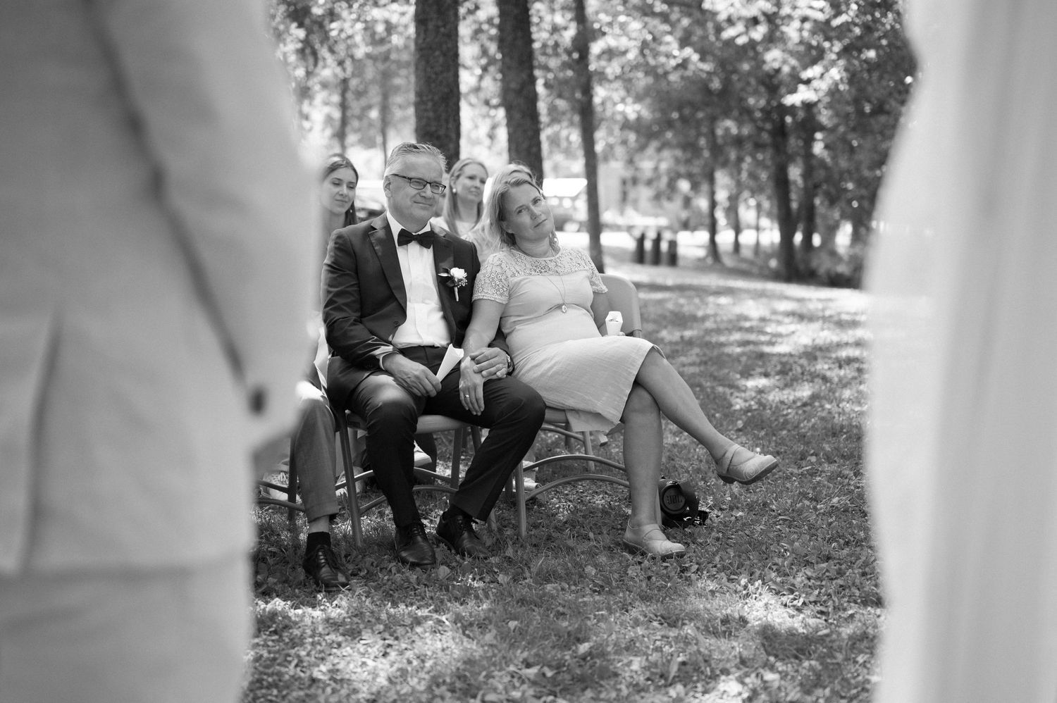Outdoor wedding ceremony setup with white columns and guests seated on white chairs in a forest setting.
