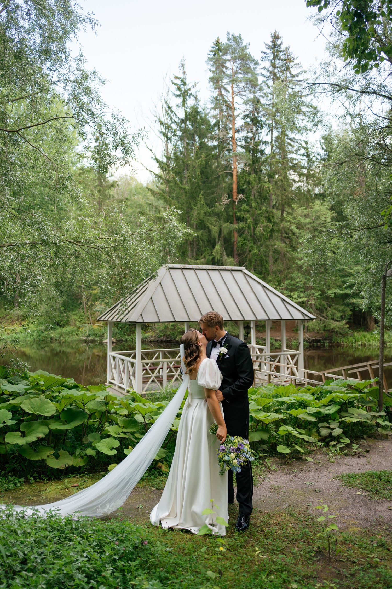 A wedding couple poses by a charming gazebo surrounded by towering evergreens and lush vegetation.