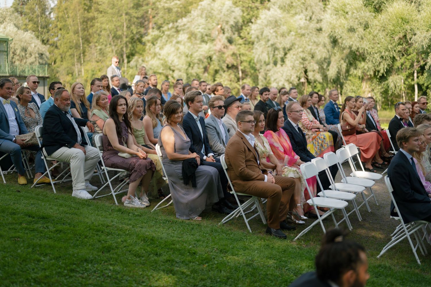 Wedding guests seated in white chairs on a lawn watch the outdoor ceremony.
