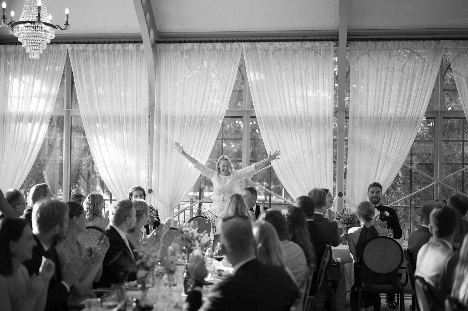 Panoramic view of wedding guests celebrating in a bright reception hall with white curtains.