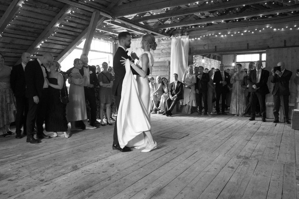 Black and white photo of couple's first dance in a rustic barn with string lights overhead.