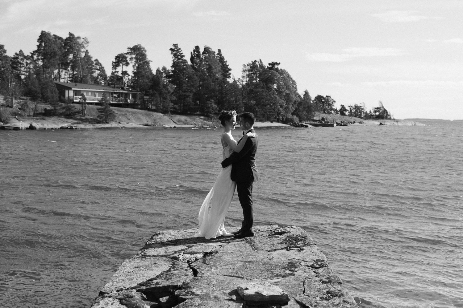 A black and white wedding photo of a couple standing on rocky shoreline overlooking water.