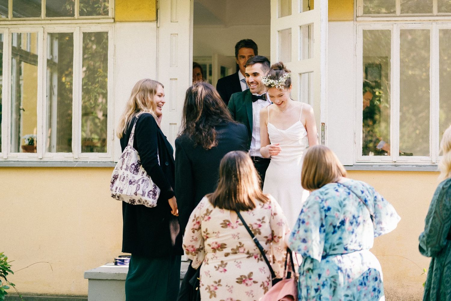 Wedding guests in floral dresses gather outside a yellow building with white trim.