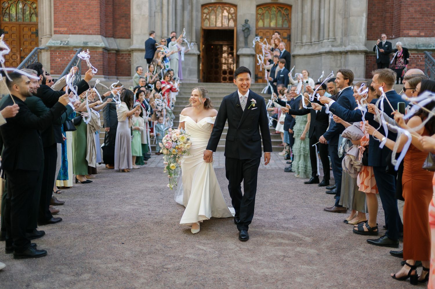 Newlywed couple walks through cheering guests waving white flags outside a historic brick building.