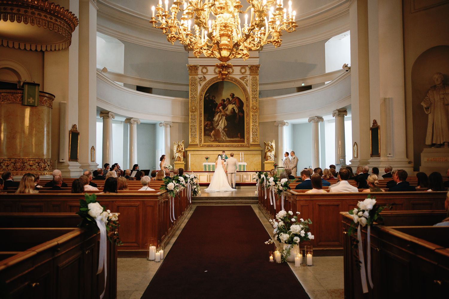 Elegant church wedding ceremony with floral-lined aisle, wooden pews, grand chandelier, and ornate altar painting.