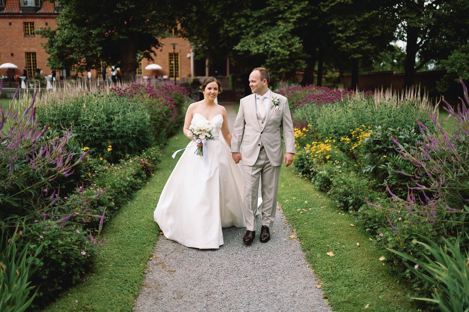 Bride and groom walk hand in hand through a lush garden path lined with colorful wildflowers on their wedding day.