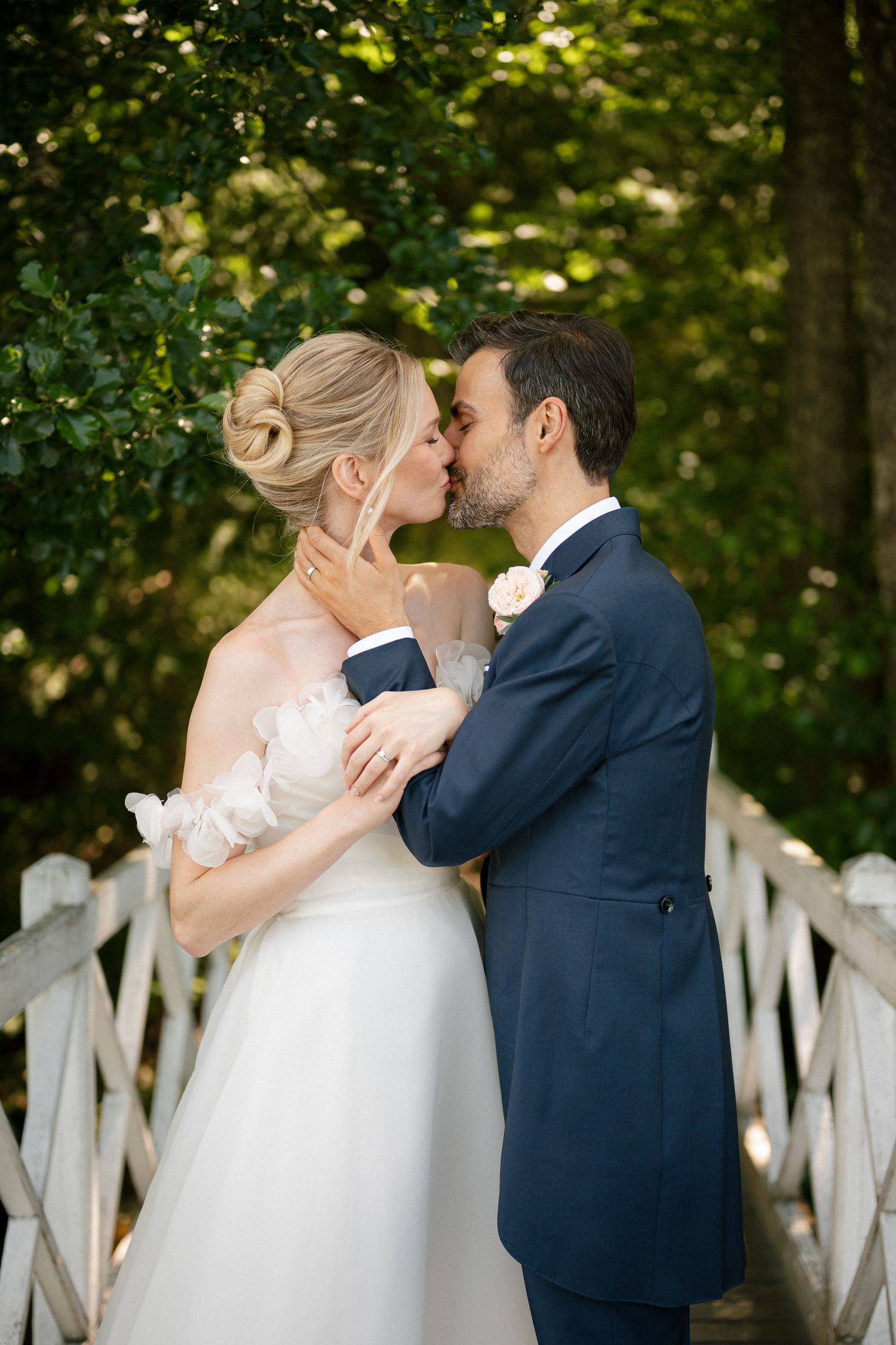 A bride and groom share a romantic kiss on a white bridge surrounded by lush green trees on their wedding day.