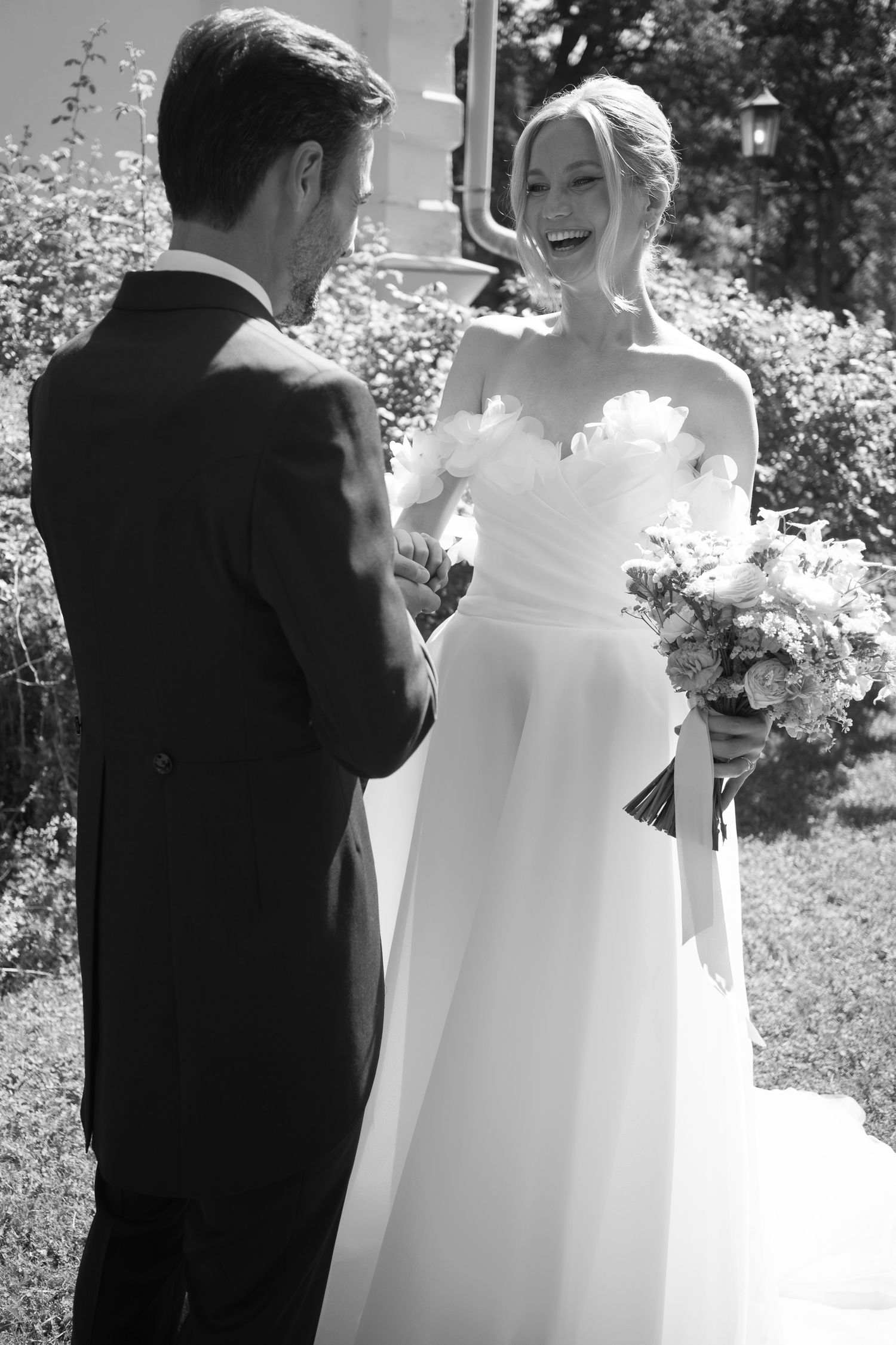 Black and white photo of a smiling bride in a strapless gown holding a bouquet, facing her groom in a suit outdoors.