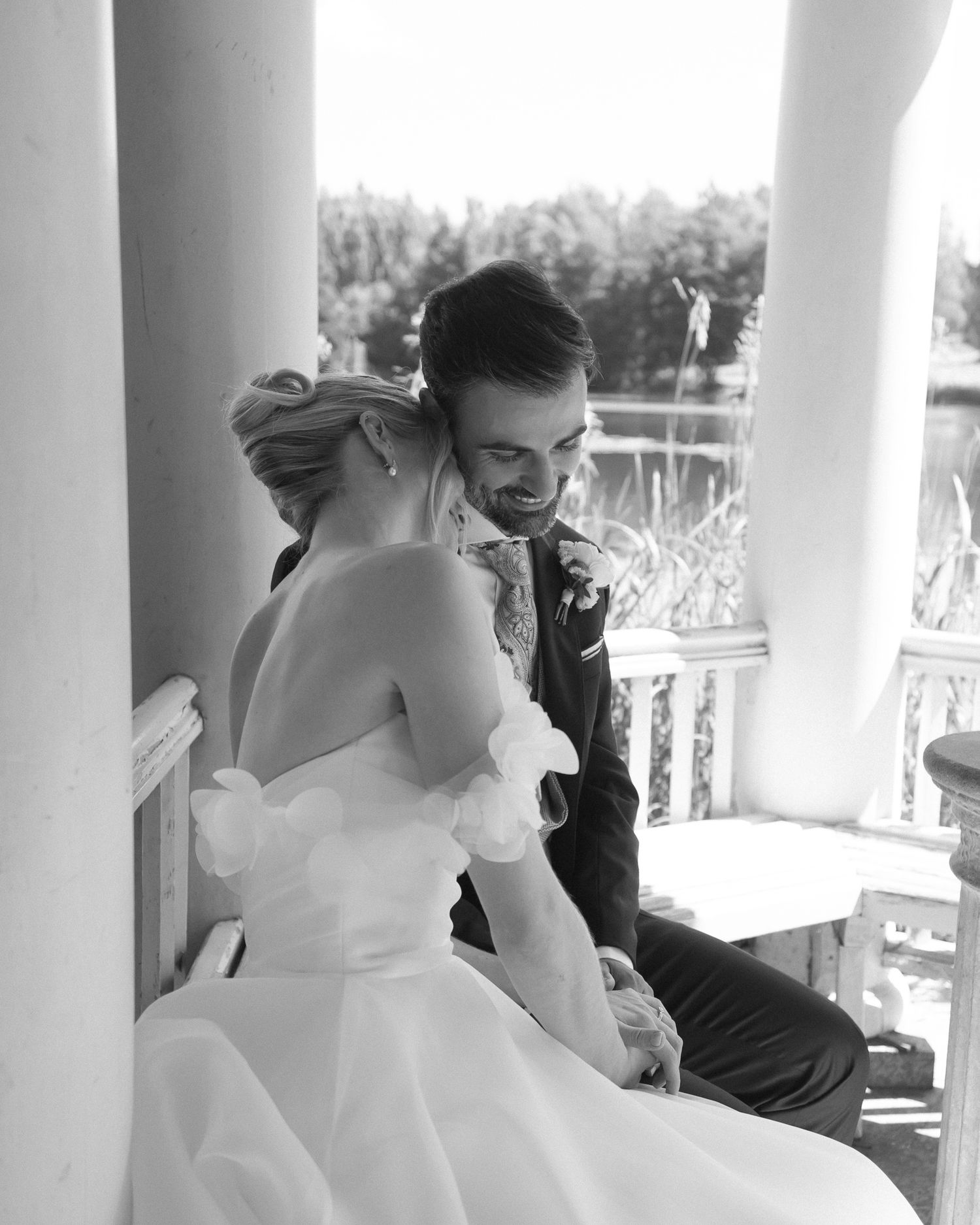 Black and white wedding photo of bride and groom sharing an intimate moment on a balcony overlooking water.