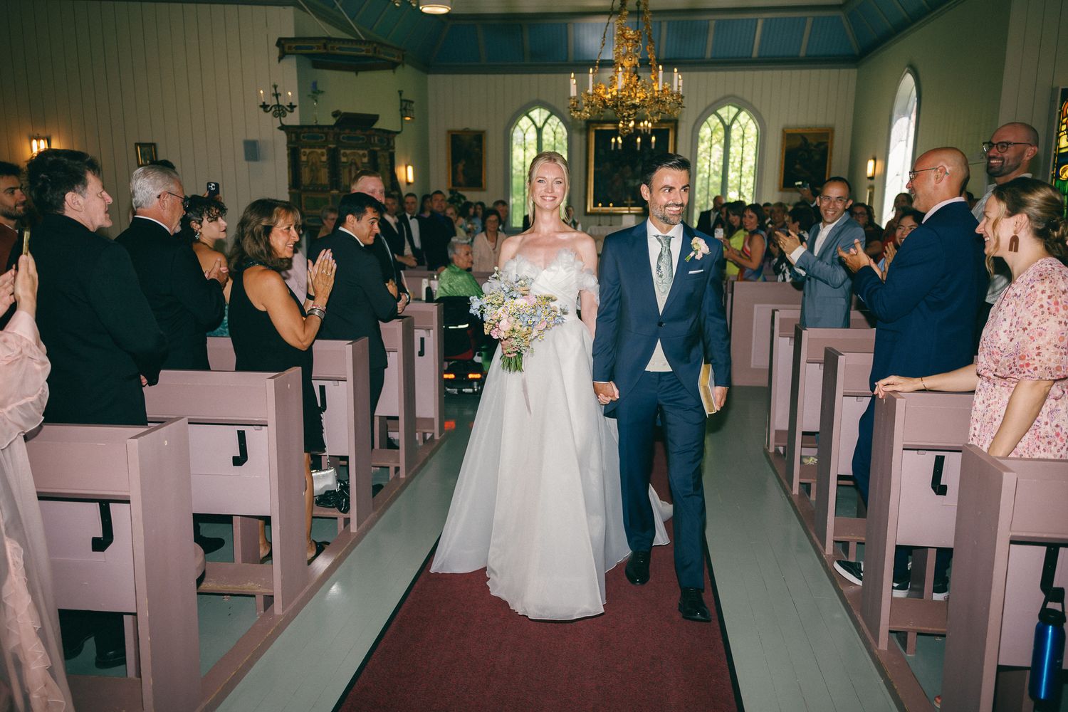 Bride and groom walk down church aisle after wedding ceremony as guests applaud in wooden pew chapel.