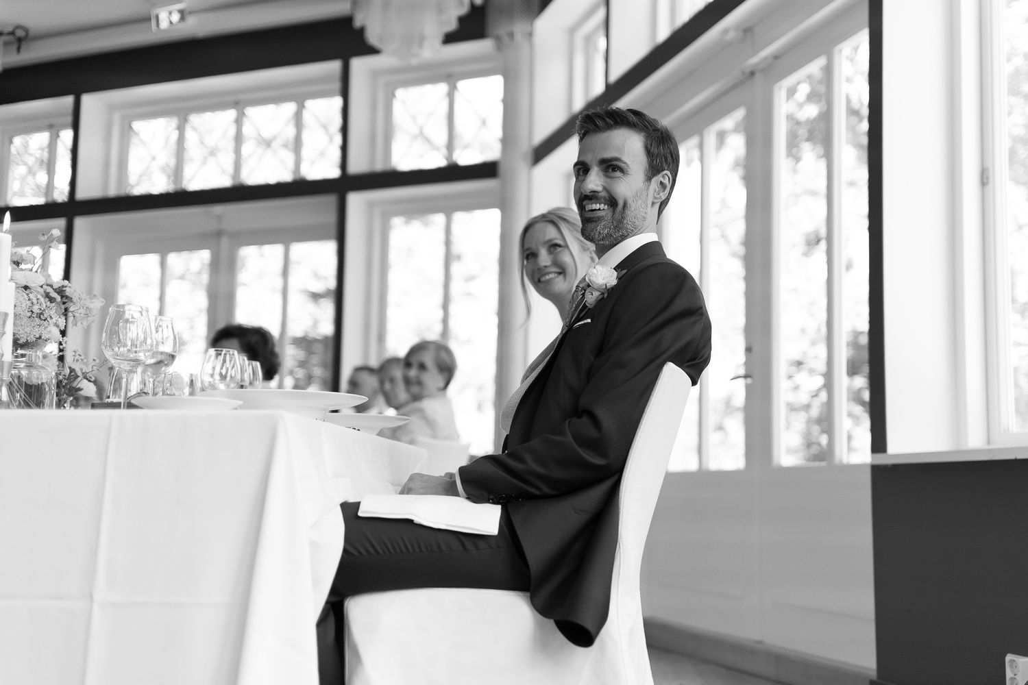Black and white photo of a smiling groom in a suit sitting at a wedding reception table with guests in the background.