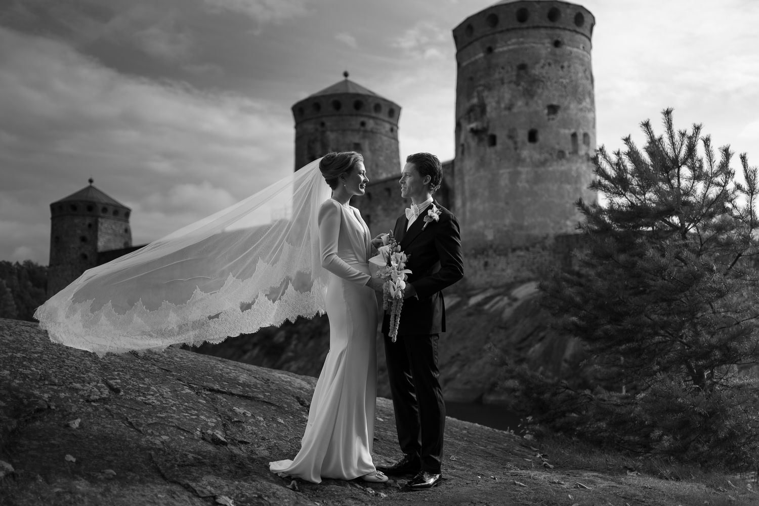 Black and white wedding photo of bride and groom standing before historic castle towers, veil flowing in wind.