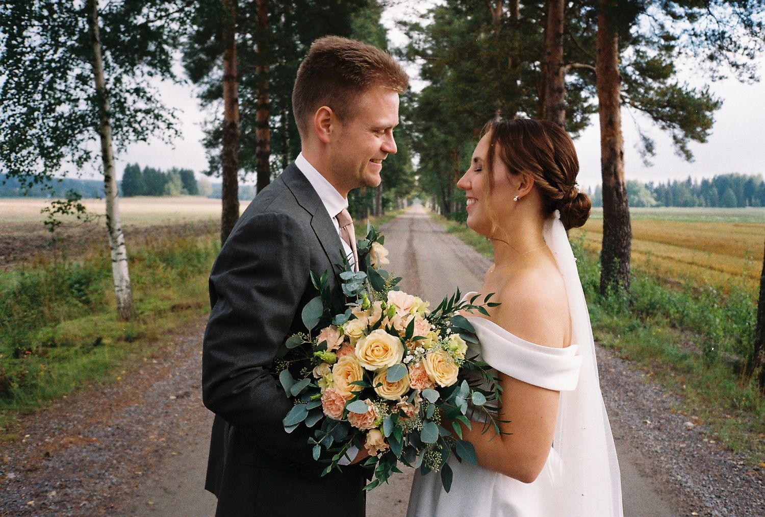 Elegant wedding couple poses on a tree-lined path with peach roses in their bouquet.