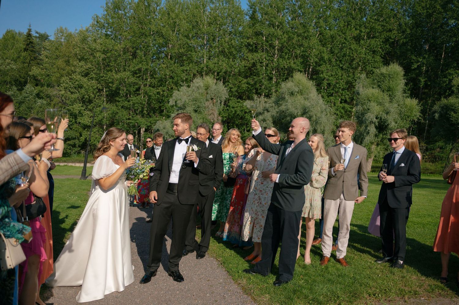 Wedding guests gather outdoors on a path surrounded by lush green trees during a summer celebration.