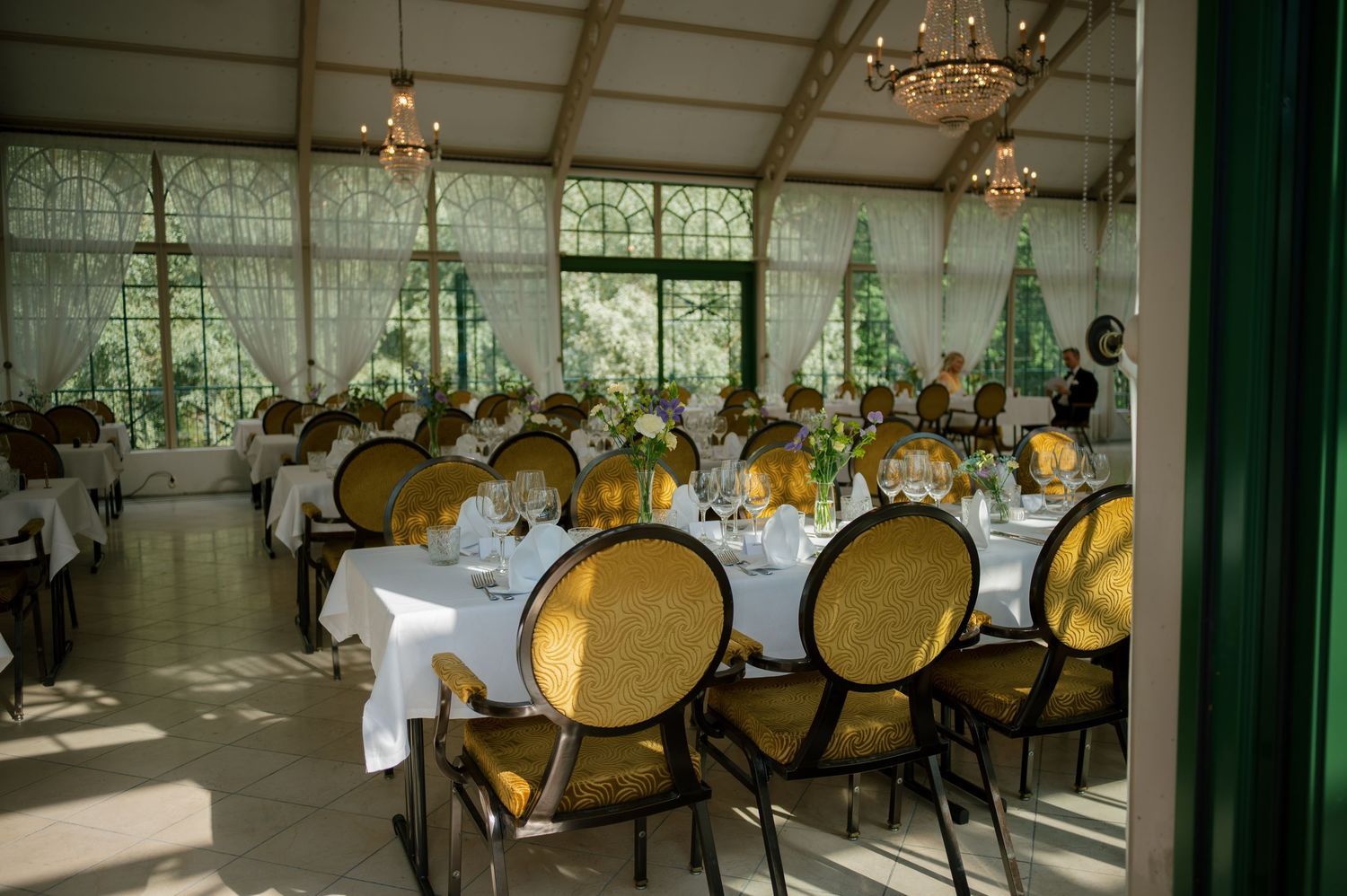 Elegant dining room with chandeliers, gold-backed chairs, and white tablecloths in natural light.