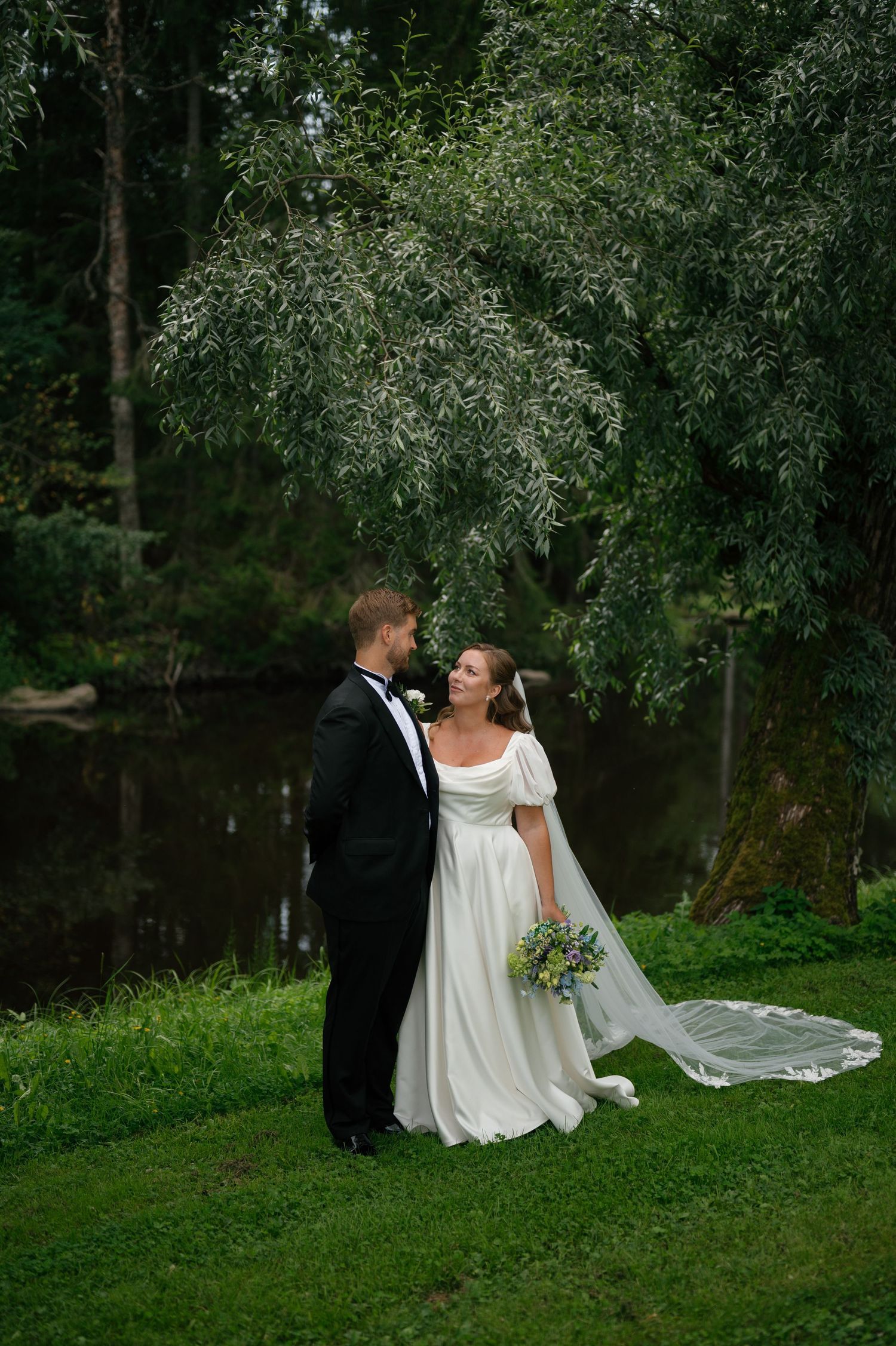 Wedding couple poses on grassy lawn near water with trees in background.