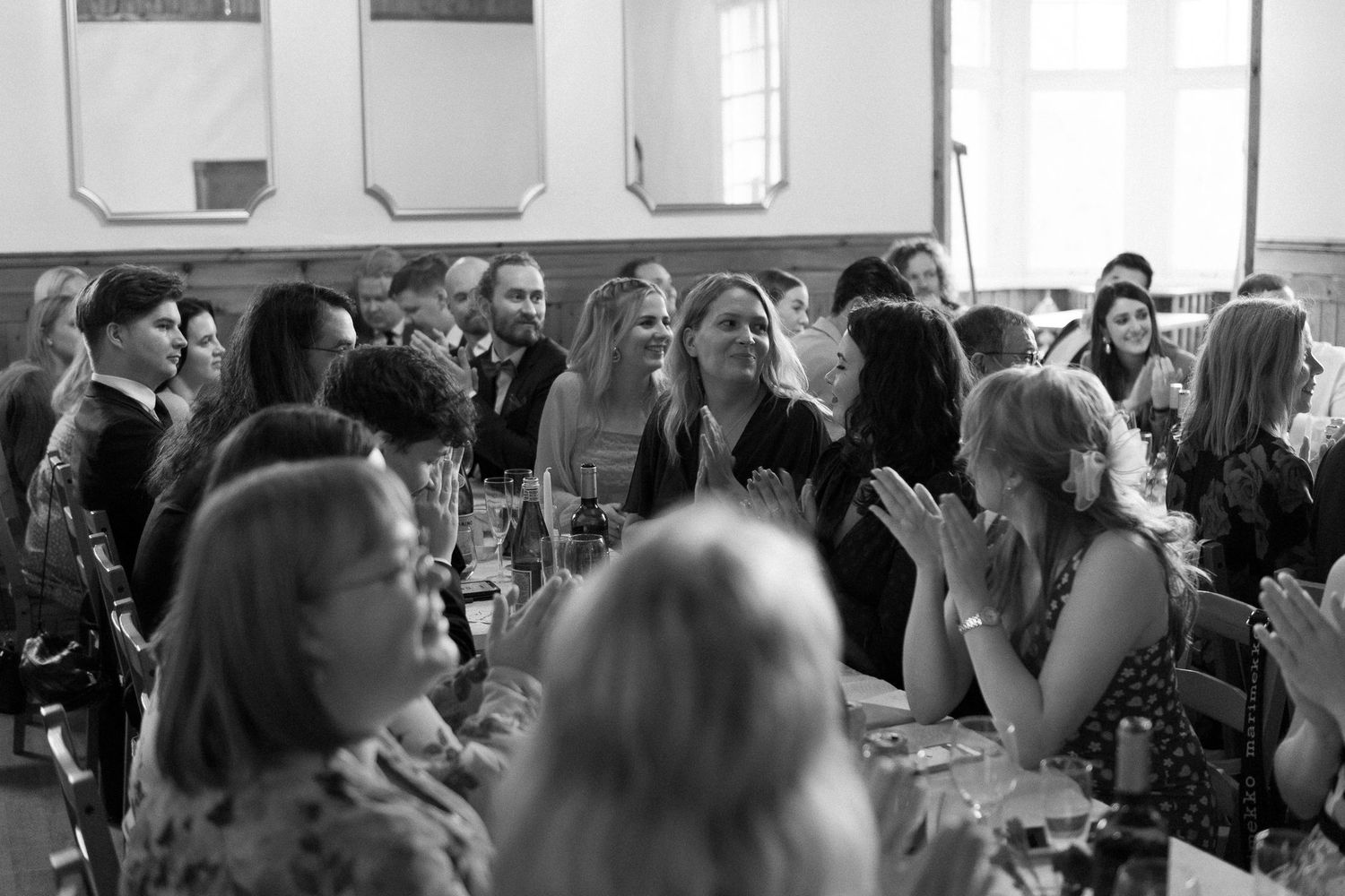 Black and white image of a lively wedding reception with guests seated at tables.