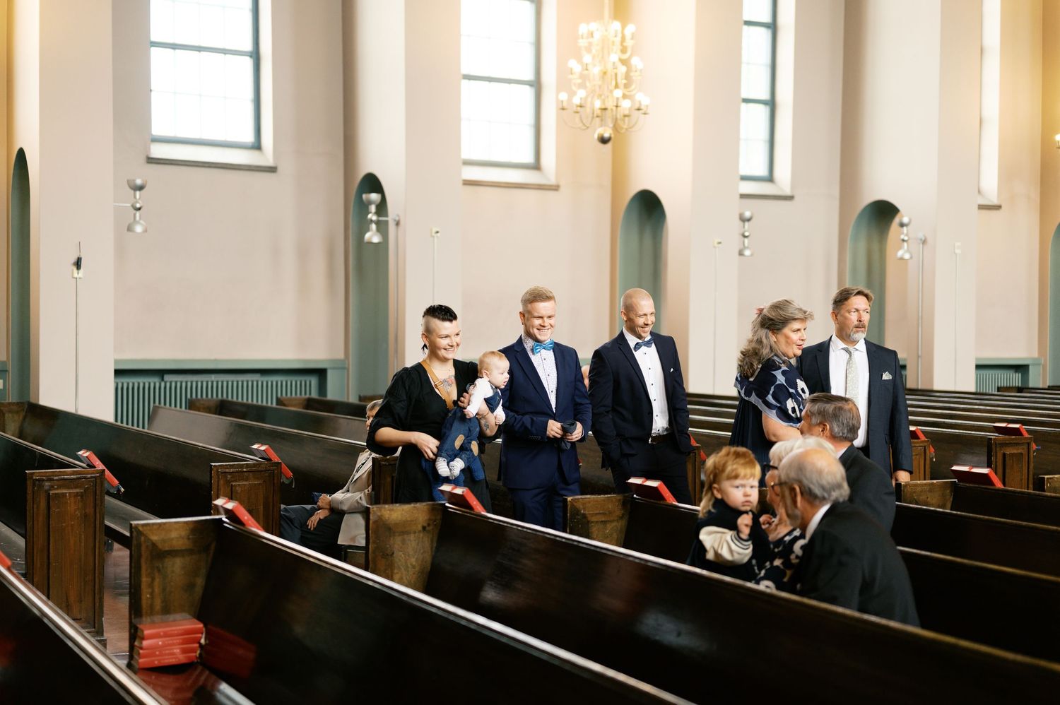 A panoramic view of people seated in church pews during a formal ceremony in a bright, white-walled sanctuary.