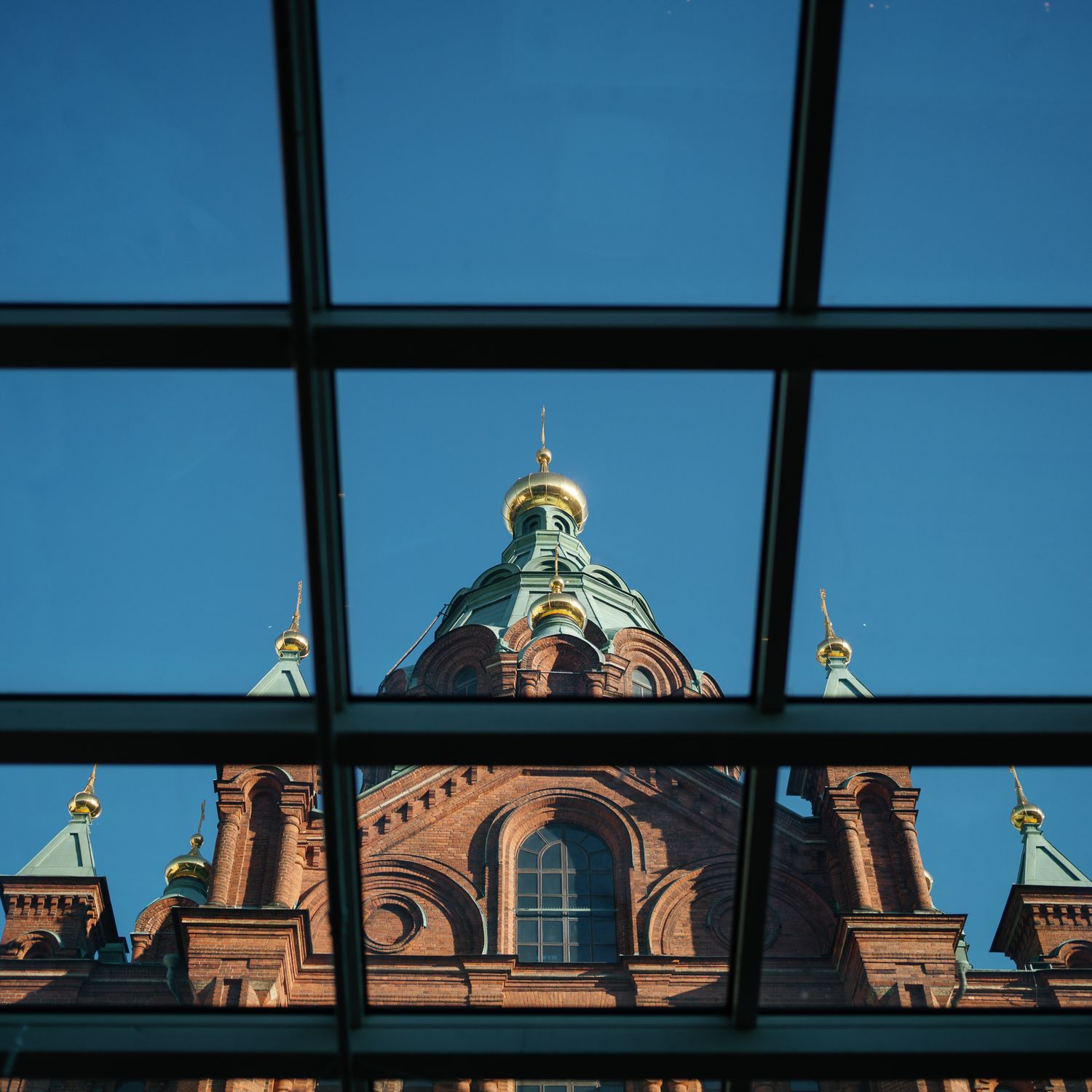 Red brick Orthodox church dome with golden cupola viewed through modern glass ceiling against blue sky.
