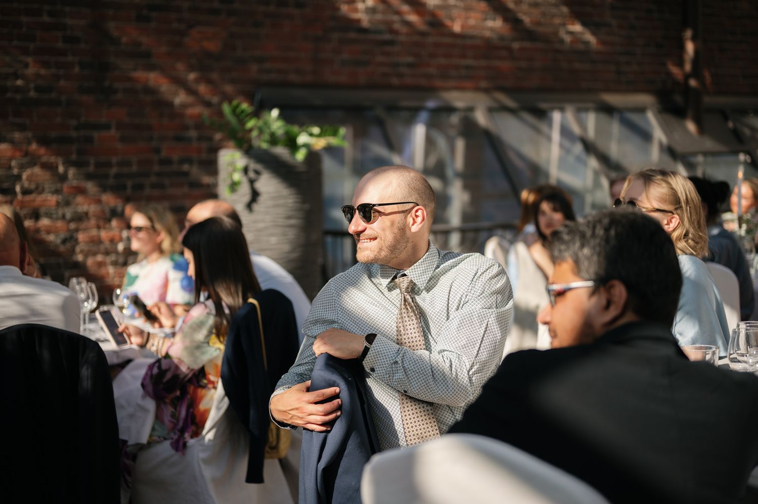 Bald man in sunglasses and gray suit smiling at an outdoor wedding reception with guests seated nearby.