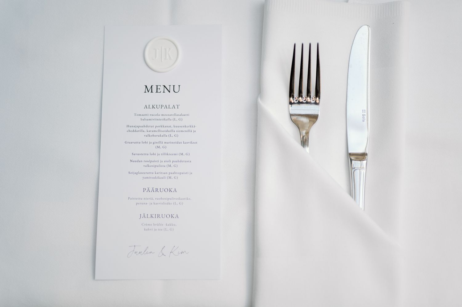 Elegant wedding menu card with wax seal beside folded white napkin with silver fork and knife on white tablecloth.