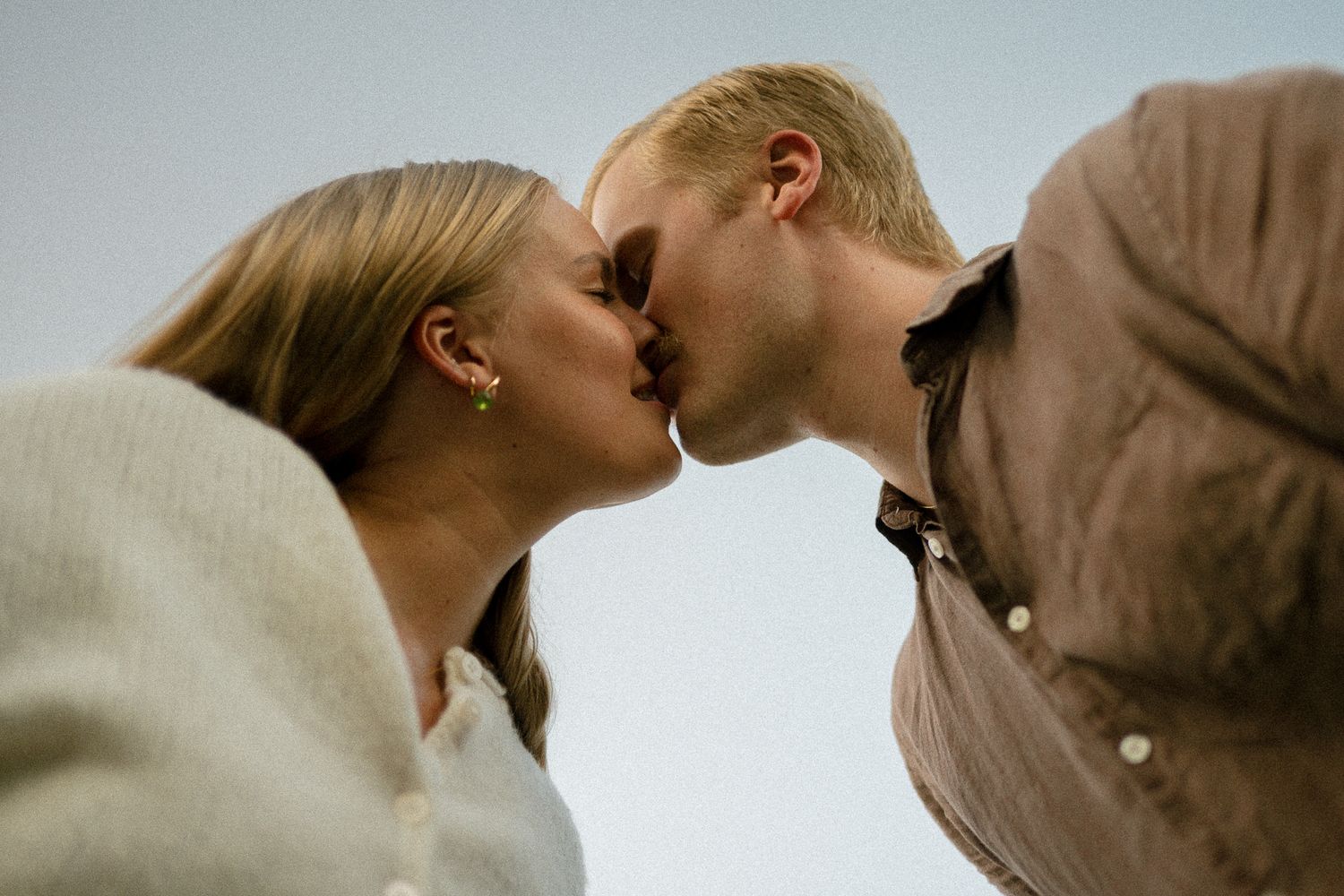 A couple shares a tender kiss outdoors, photographed from below against a soft gray sky.