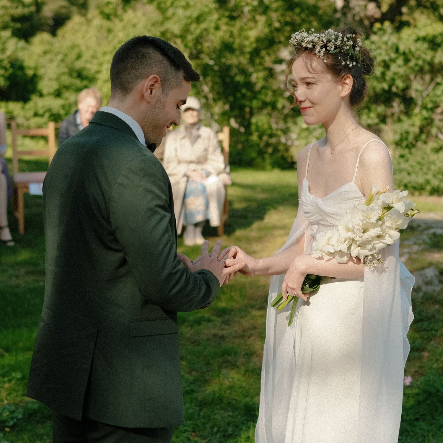Groom placing wedding ring on bride's finger outdoors, bride holding white bouquet wearing floral crown.