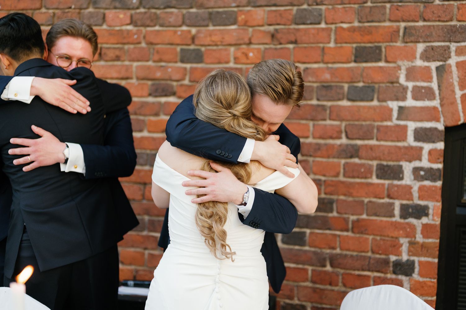 Bride and groom sharing heartfelt hugs with wedding guests against a rustic brick wall backdrop.