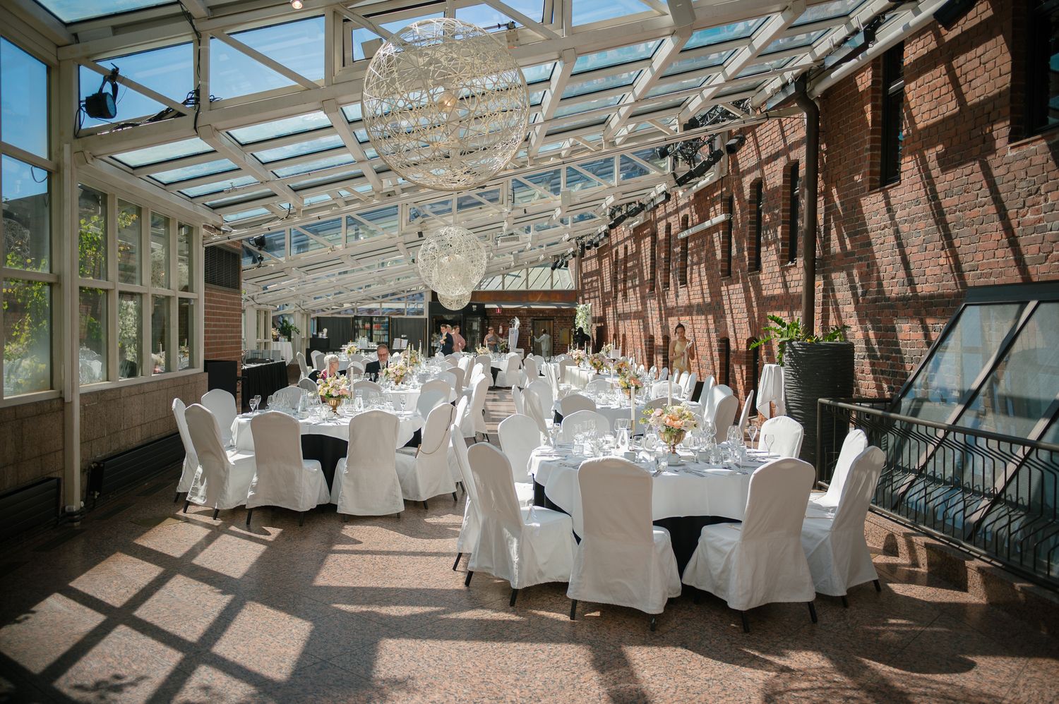 Elegant wedding reception in a glass-roofed conservatory with white draped chairs, round tables, and a chandelier centerpiece.