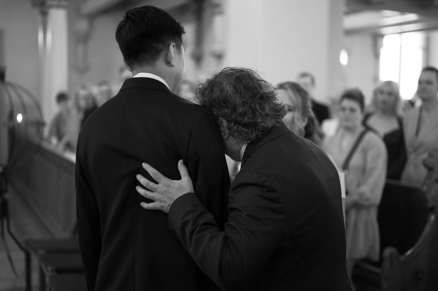 Two men in suits share an emotional embrace inside a church during a formal ceremony.