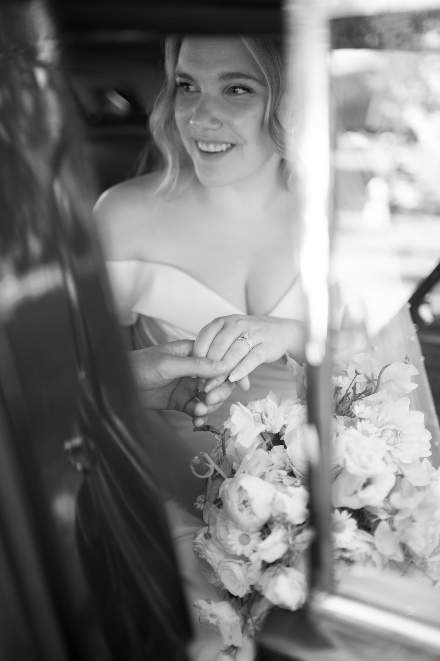 Black and white photo of a smiling bride in a strapless gown holding a floral bouquet, seated in a vintage car.