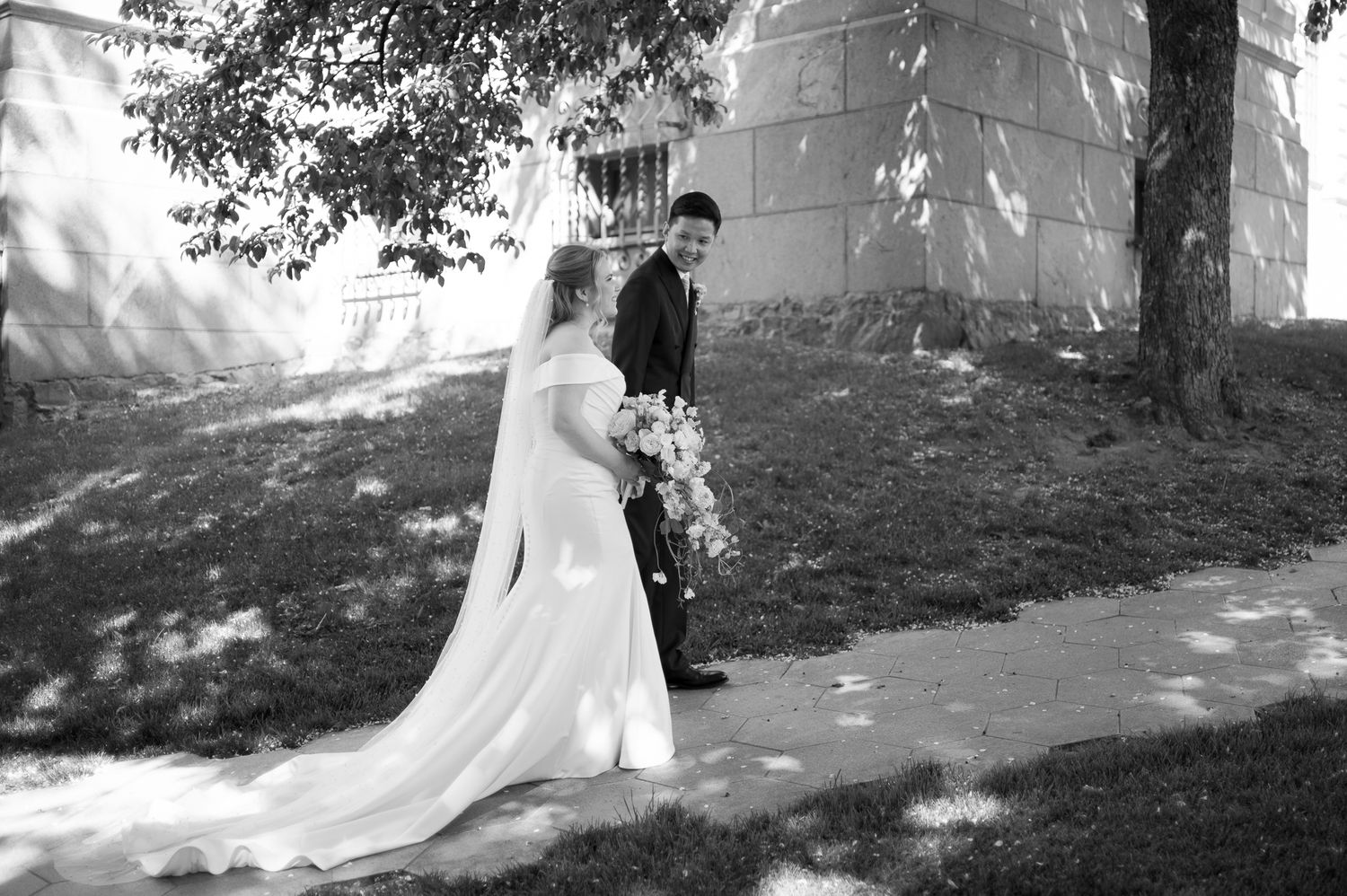 Black and white photo of a bride in a flowing gown and veil holding a bouquet, standing with a groom outdoors.