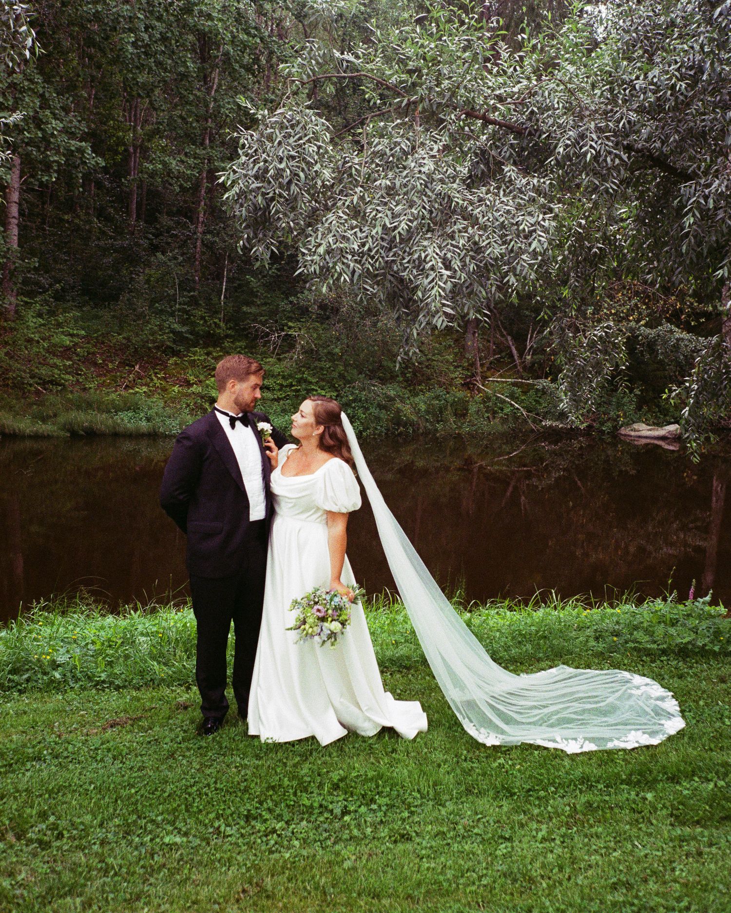 A bride and groom share a romantic moment in a lush green garden near a pond, she wearing a flowing white gown with veil.