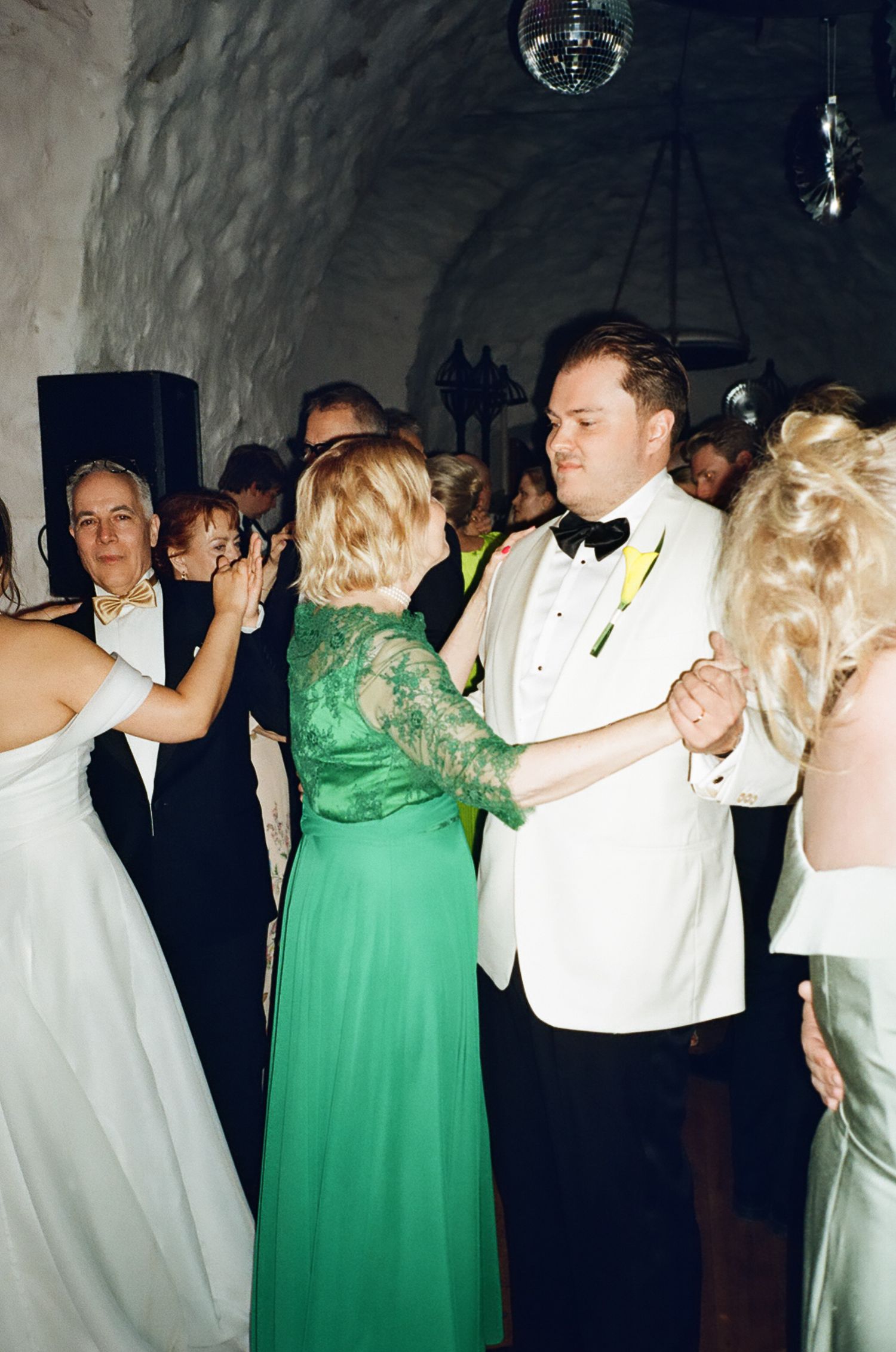 Guests dancing at a wedding reception, featuring a woman in an emerald green gown and a man in a white tuxedo.