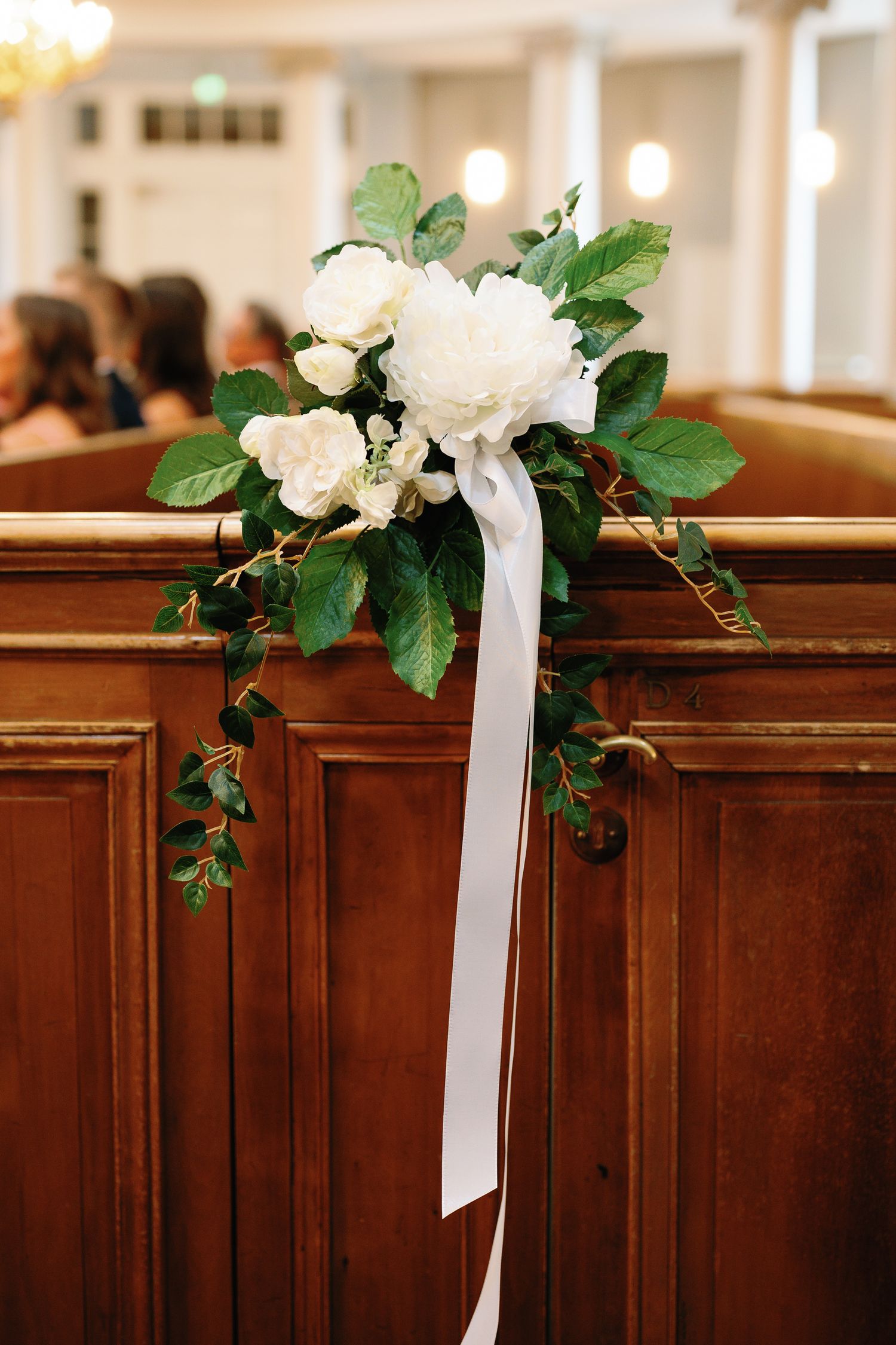 White floral bouquet with green leaves and white ribbon tied to wooden church pew at elegant wedding ceremony.