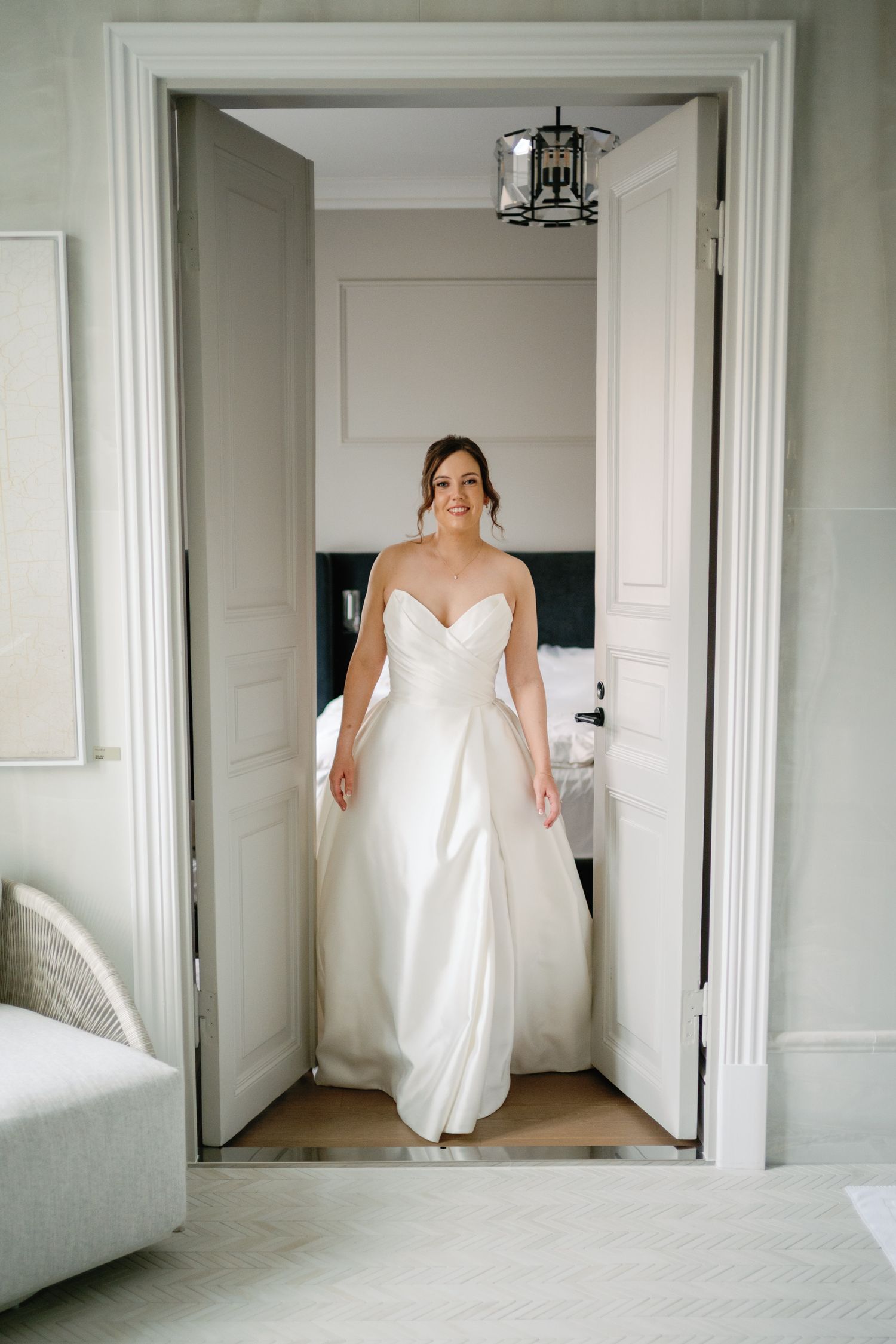 Bride in strapless white satin ball gown standing in elegant doorway of bridal suite, smiling softly.